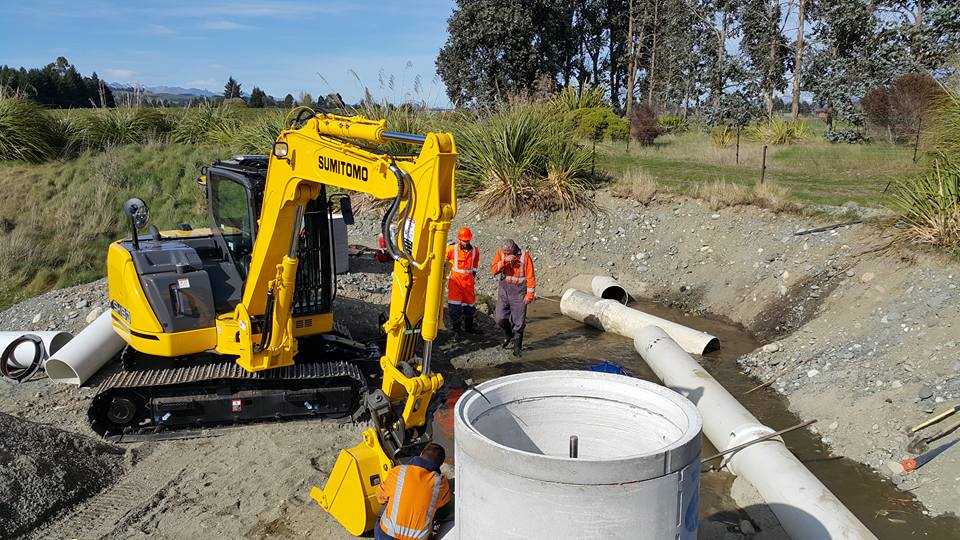 A tank and some pipes waiting to be assembled in an excavated area. Three Te Anau Earthworks staff are attending to a yellow Sumitomo digger, ready to start work. 