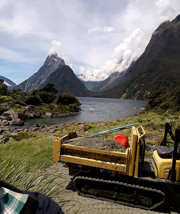 A yellow Yanmar C12R-C near the outlet of the Cleddau river, Milford Sound with it's deck full with gravel. Mitre Peak can be seen in the background.