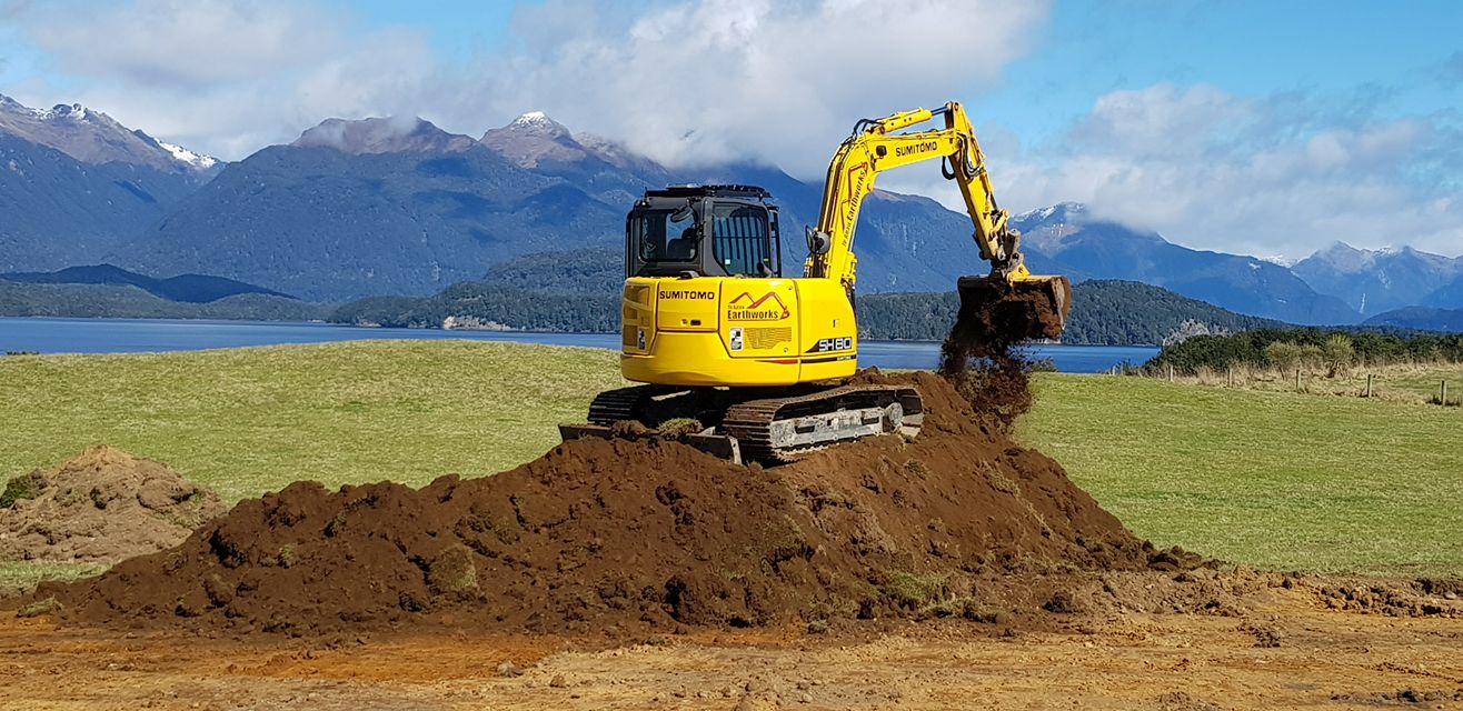 Large yellow Sumitomo digger perched on a hill of dirt while it's digging. Lake Te Anau and the Murchison mountains can be seen in the background.