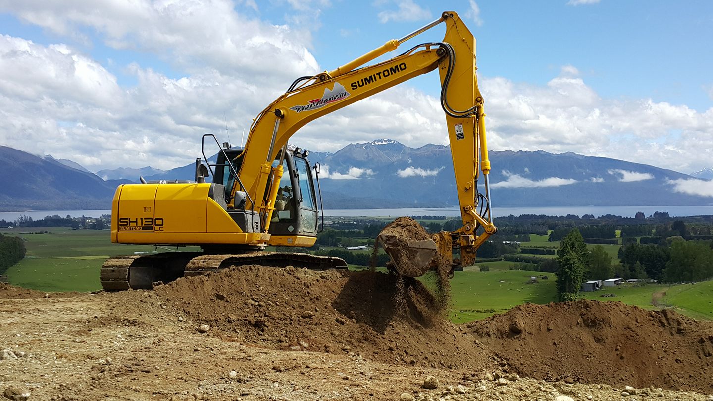 Large yellow Sumitomo digger perched on a hill while it's digging. Lake Te Anau, Te Anau township and the Murchison mountains can be seen in the background.