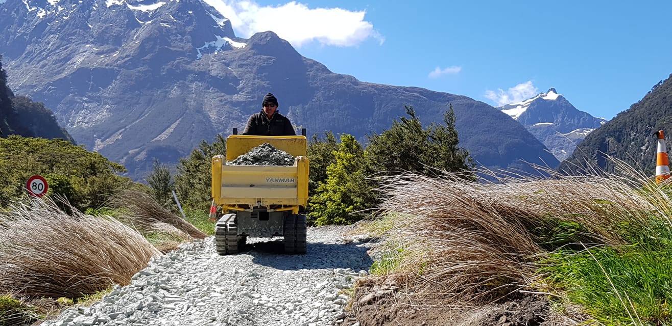 A yellow Yanmar C12R-C and driver working on a narrow gravel track with large mountains looming in the background. 