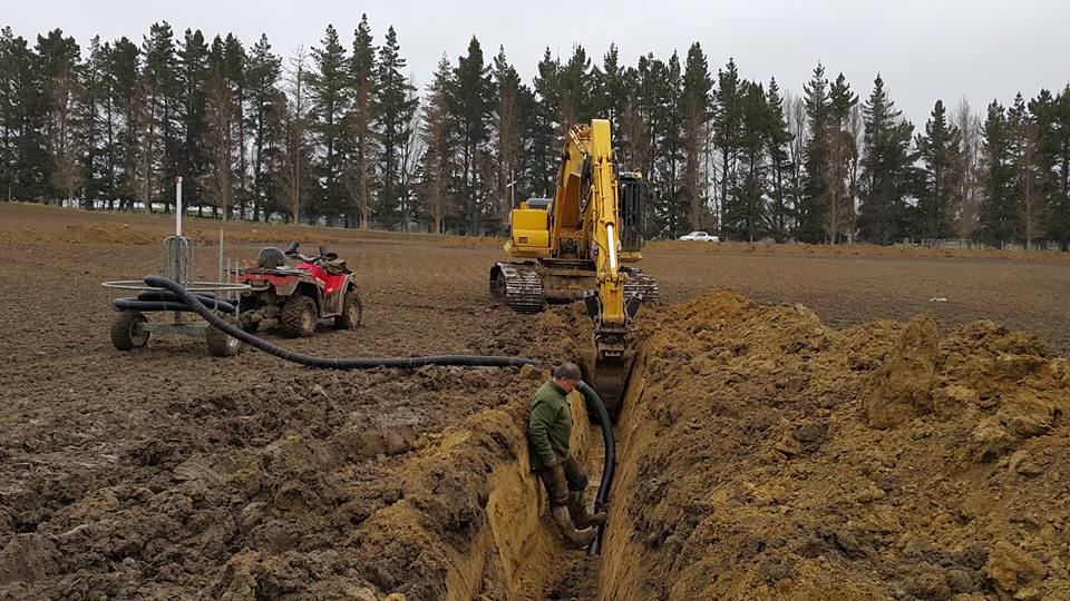 A yellow digger creating a ditch in a muddy paddock. There is a worker standing in the ditch with a pipe which leads back to his four-wheeler motorbike and a trailer with a coil of pipe.