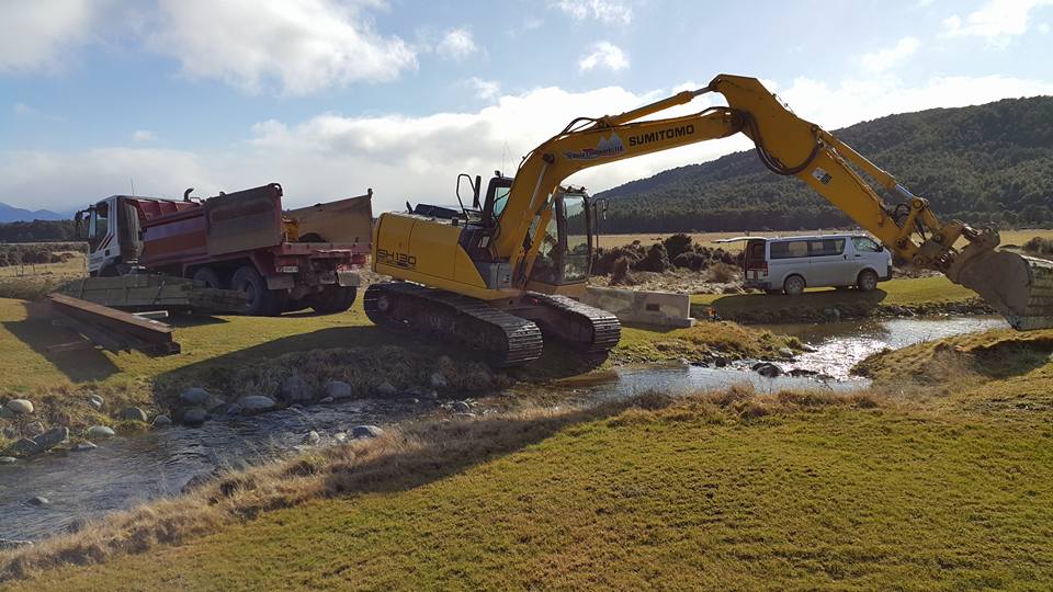 A large yellow sumitomo digger about to cross a small stream. More equipment is waiting by a truck and a white van in the background.